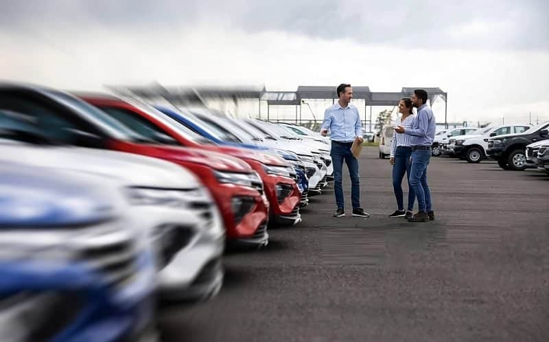 People discussing near rows of parked cars in a dealership lot, suggesting car sales or inspection.